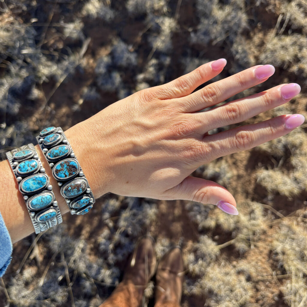 A hand with long pink nails is shown wearing two chunky turquoise and silver bracelets. The background is dry grass, and brown boots are visible at the bottom of the image.