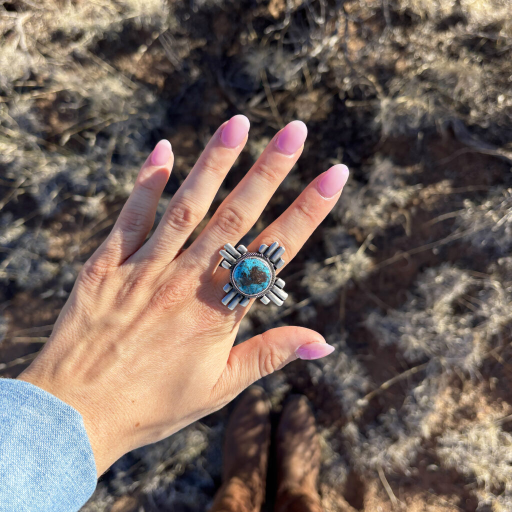 A hand with pink nails wearing a large turquoise and silver ring is held out over dry grass, with sunlight casting shadows and the persons boots visible at the bottom of the image.