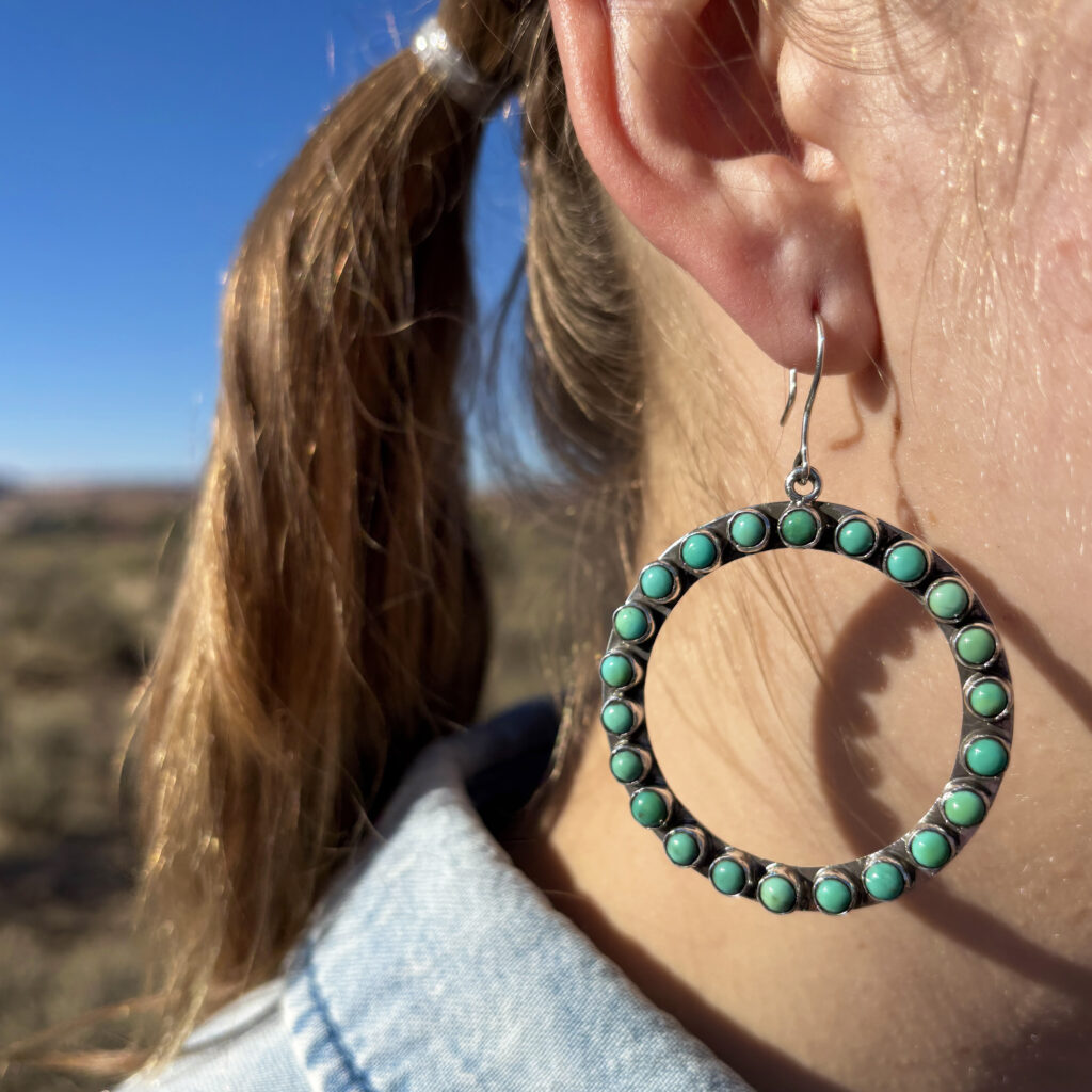 A close-up of a person’s ear wearing a large circular earring decorated with turquoise stones. The person has light brown hair in a ponytail and is standing outdoors under a clear blue sky.