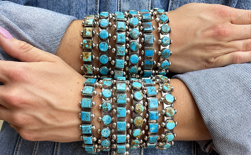 A person wearing a denim shirt crosses their arms, displaying multiple chunky silver bracelets adorned with turquoise and stone inlays in various shapes and shades of blue and green.