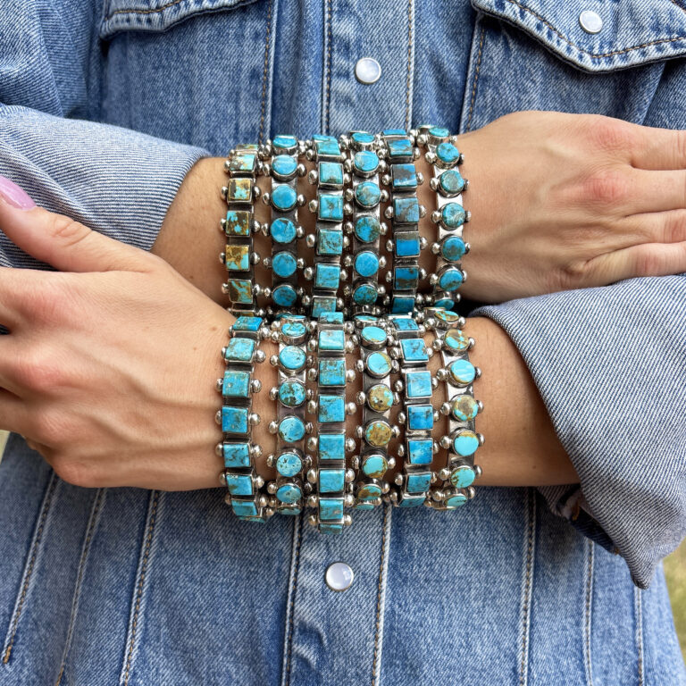 A person wearing a denim jacket crosses their arms, displaying multiple turquoise and silver bracelets stacked on both wrists. The bracelets feature round and square turquoise stones.