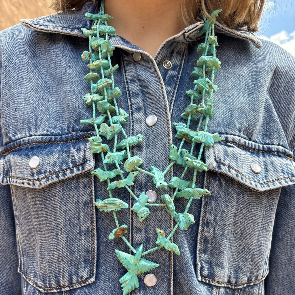 A person wearing a blue denim jacket is shown from the shoulders down, wearing layered turquoise necklaces featuring multiple animal-shaped beads. The sky is visible in the background.