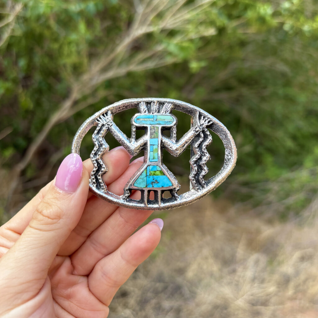 A hand with pink-painted nails holds an oval silver and turquoise belt buckle featuring a stylized, geometric figure and zigzag patterns, with green foliage in the blurred background.