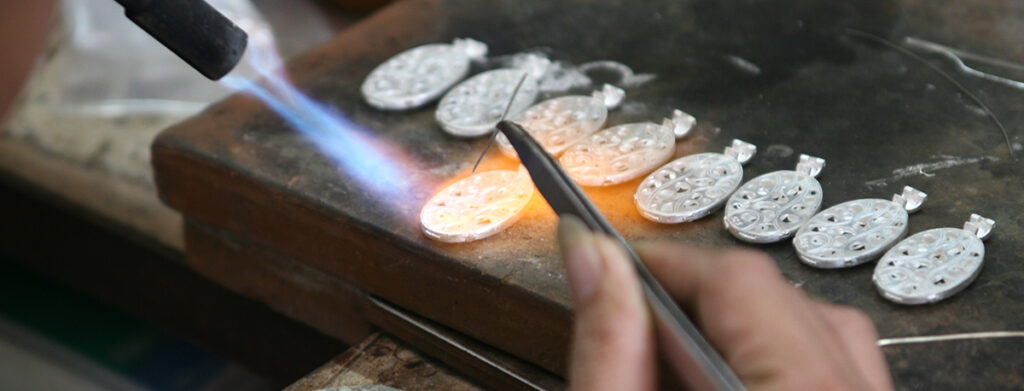 A person uses a torch and tweezers to work on small, oval-shaped metal pieces arranged in a row on a workbench, possibly making jewelry or metal crafts.