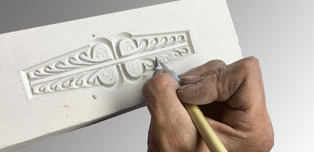 A close-up of a person’s hands carving an intricate decorative pattern into a white rectangular piece of material using a pointed sculpting tool.