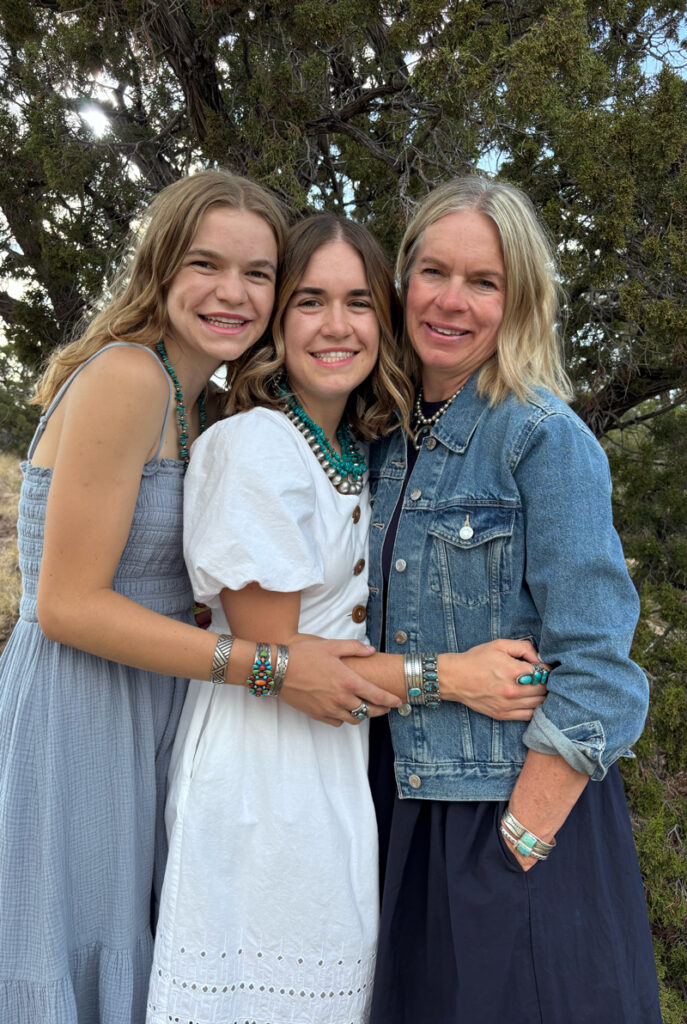Three women, likely family, stand close together outdoors, smiling at the camera. They wear casual dresses and turquoise jewelry, with trees in the background. The mood is warm and affectionate.