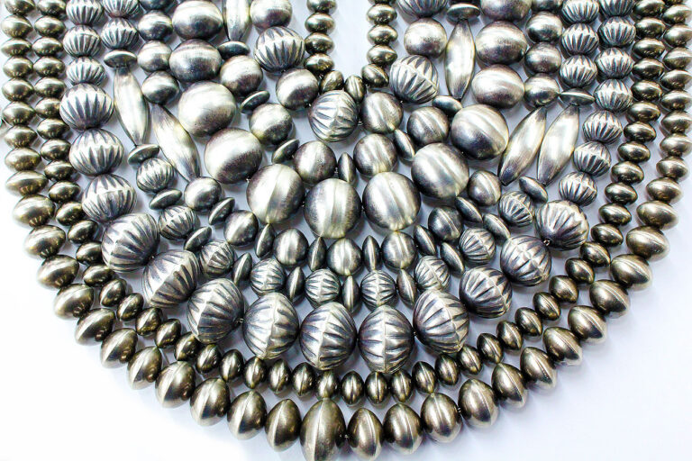 Several strands of silver beads with various shapes and textured patterns are layered on a white background, arranged in a semi-circular, overlapping display.