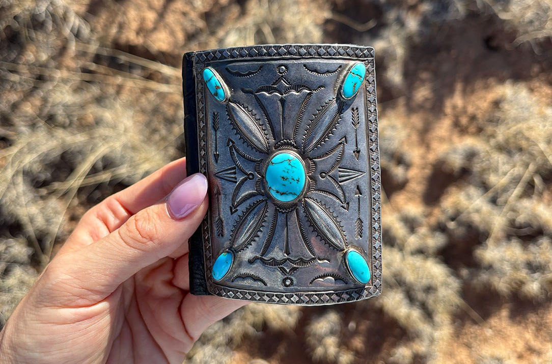 A hand with light pink nail polish holds a rectangular, ornate silver object with engraved patterns and five turquoise stones, against a blurred outdoor background with dry grass and soil.