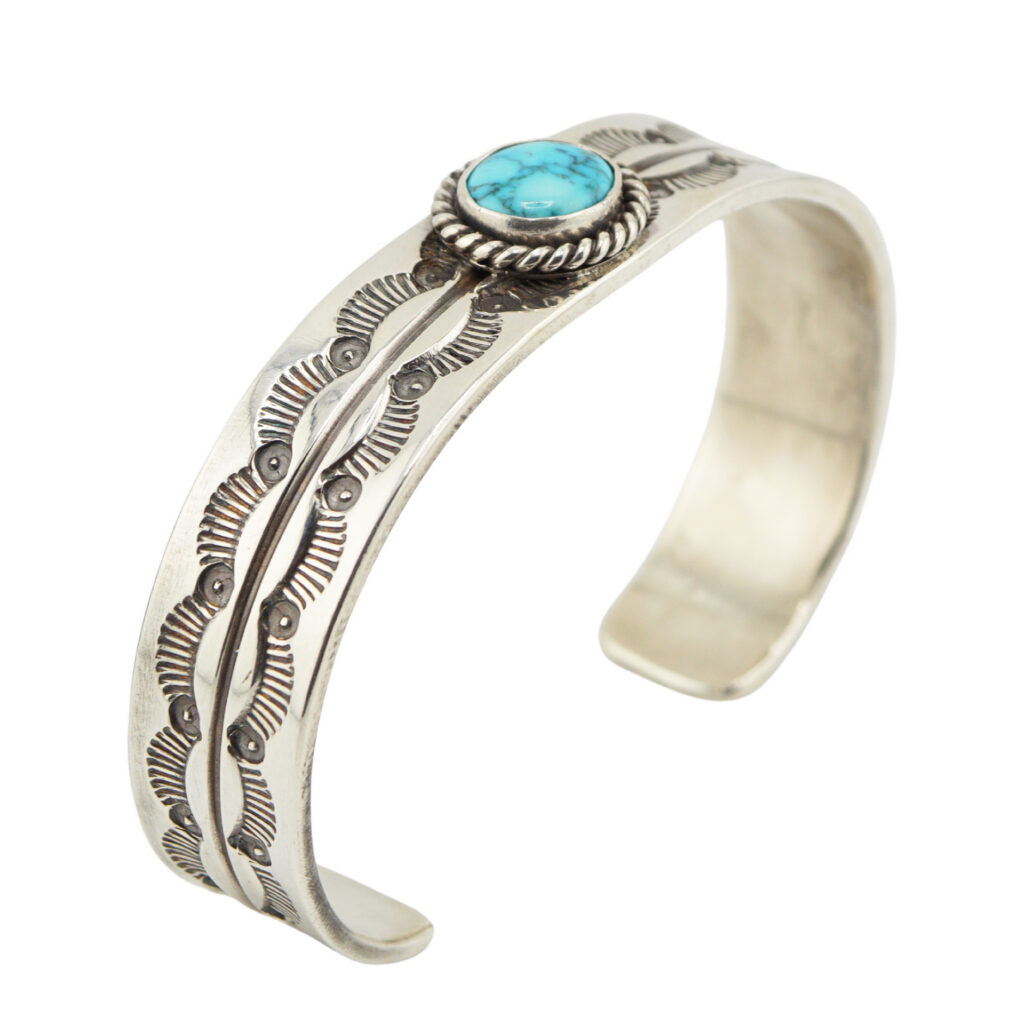 A silver cuff bracelet featuring intricate engraved patterns and a central round turquoise stone set in a twisted rope bezel.