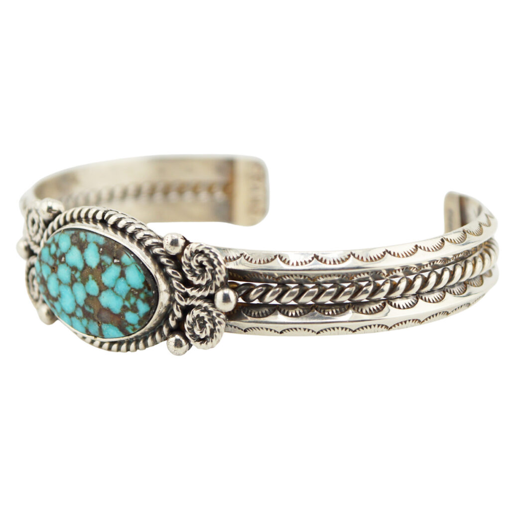 The Single Stone Spiderweb Cuff by Melvin Francis (Navajo) is a silver cuff bracelet with ornate engravings and a large oval turquoise stone featuring a webbed pattern in a decorative bezel.