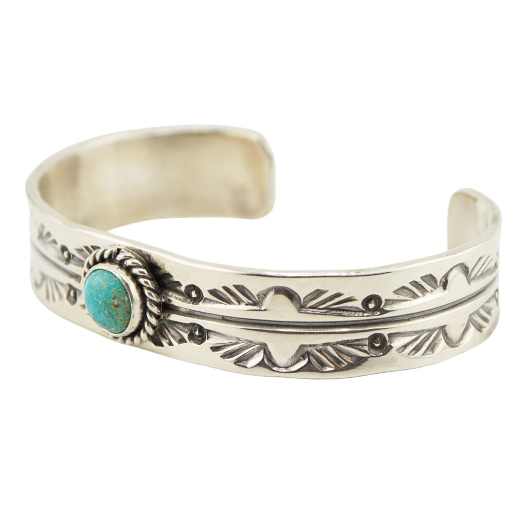 A silver cuff bracelet featuring intricate engraved patterns and a round turquoise stone set in a twisted bezel on one side.