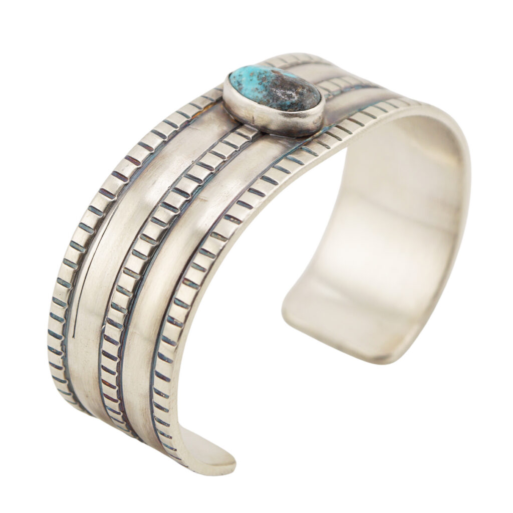 A wide silver cuff bracelet with engraved parallel lines and a single oval turquoise stone set in the center.