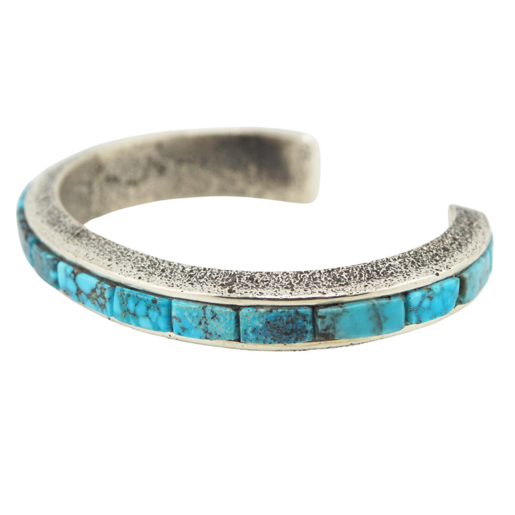 A silver cuff bracelet featuring a textured finish and a row of rectangular turquoise stones inlaid along the top. The bracelet is open-ended and has an artisanal, rustic look.