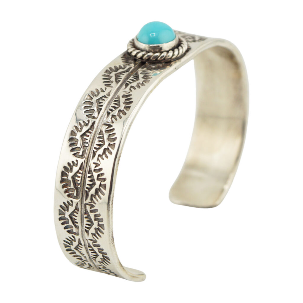 A silver cuff bracelet with intricate leaf-like engravings and a round turquoise stone set in a raised, rope-edged bezel at the center.