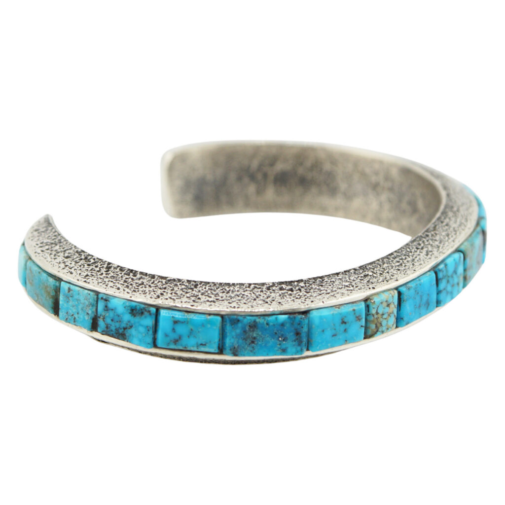 A silver cuff bracelet with a textured surface, featuring a row of square turquoise stones set along the top edge.