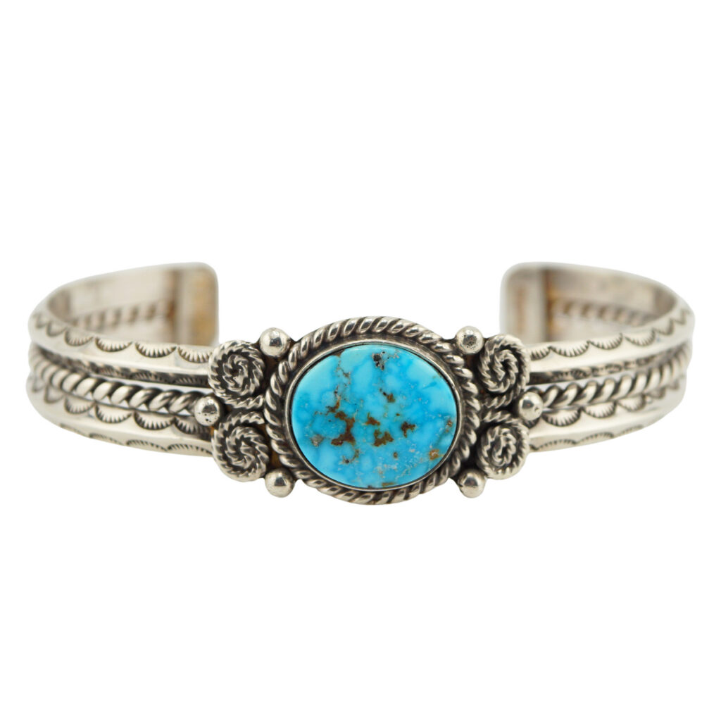 A silver cuff bracelet features intricate engraved patterns and a central oval turquoise stone set in a decorative bezel with rope and scroll details.