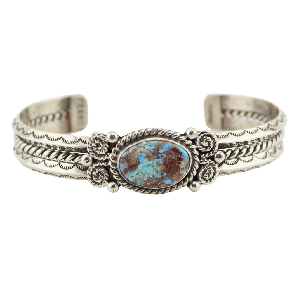A silver cuff bracelet featuring an oval turquoise stone with brown and blue patterns, detailed with intricate engraved designs and twisted rope accents on both sides.