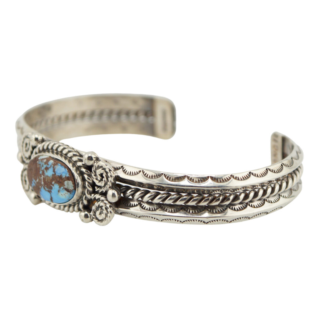 A silver cuff bracelet with intricate etched designs and twisted rope details, featuring an oval turquoise stone set in an ornate bezel on the front center.
