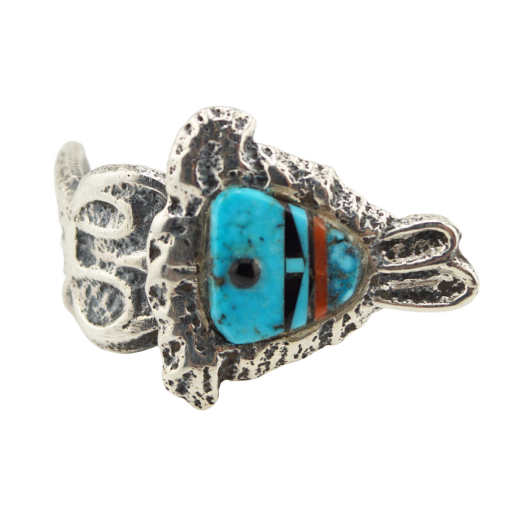 A silver ring featuring a fish-shaped design with inlaid turquoise, black, white, and brown stones forming the fishs face and body details. The band has a textured, organic pattern.