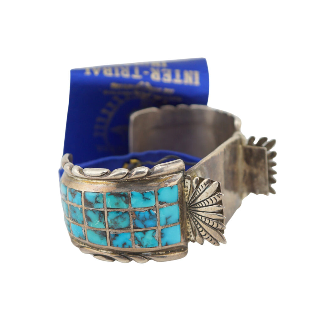A silver cuff bracelet featuring a grid of rectangular turquoise stones and intricate engraved detailing; a blue tag is attached in the background.