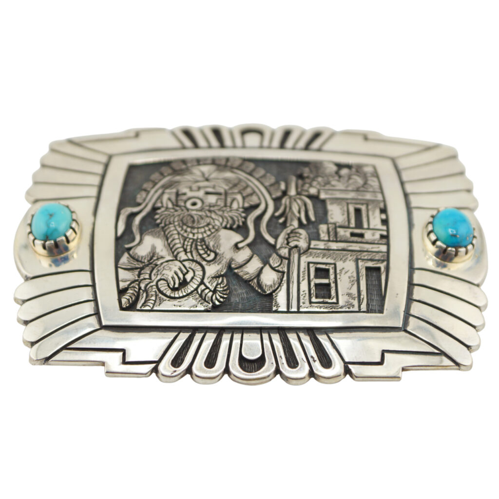 A silver belt buckle with an engraved image of a figure resembling the Hindu deity Ganesha, flanked by two turquoise stones on either side, and decorative geometric edges.